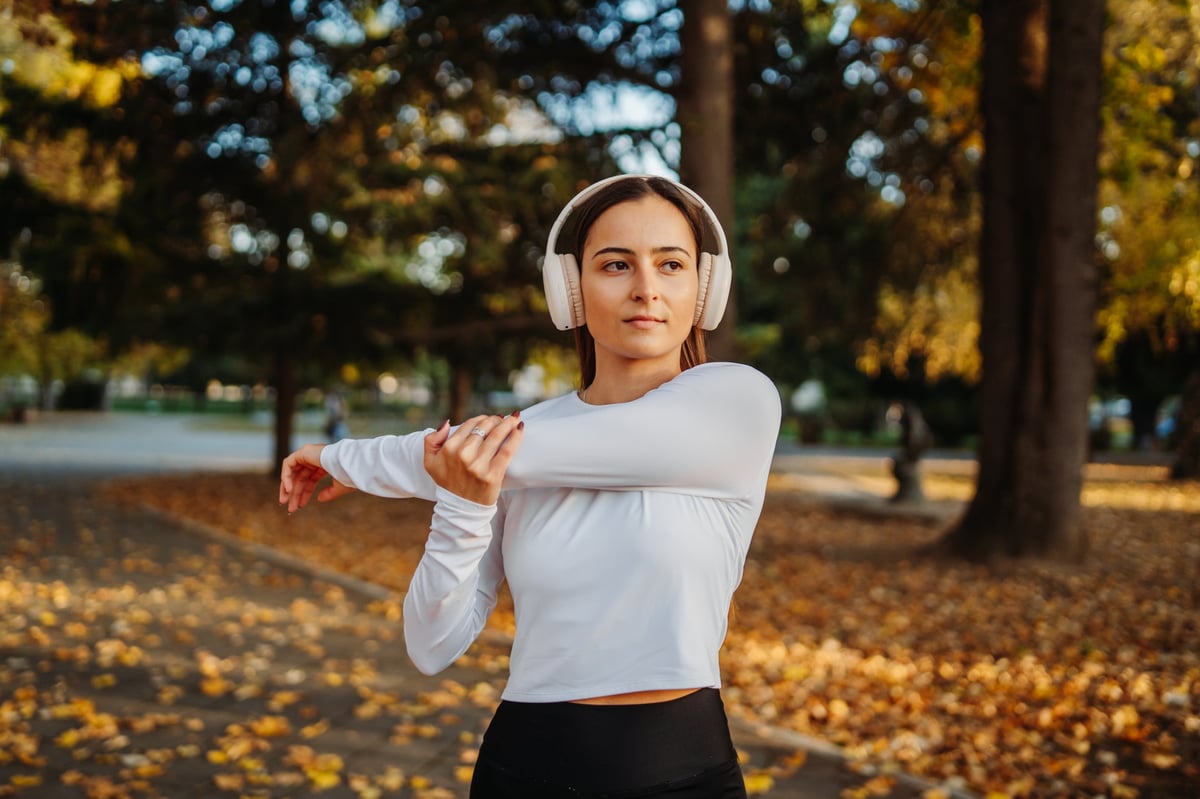 Young woman in headphones stretching her arm outside during an autumn morning workout