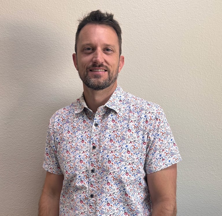 Man wearing a colorful floral short-sleeved button-up shirt smiling at camera against a plain white background